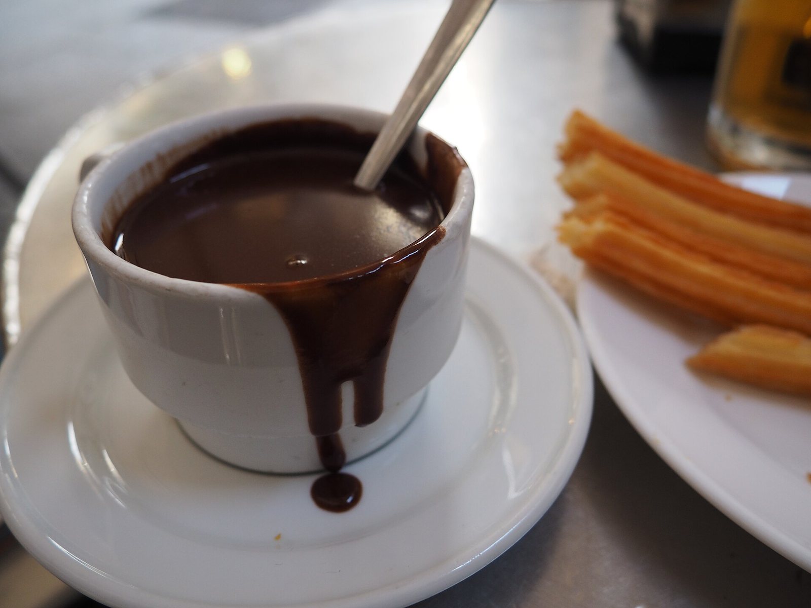 Taza de chocolate espeso con cuchara y churros al lado sobre mesa de mármol